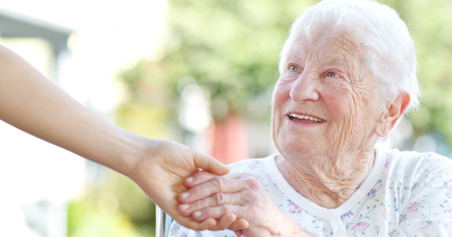 Alte Frau im Rollstuhl reicht Pflegerin die Hand und lächelt.